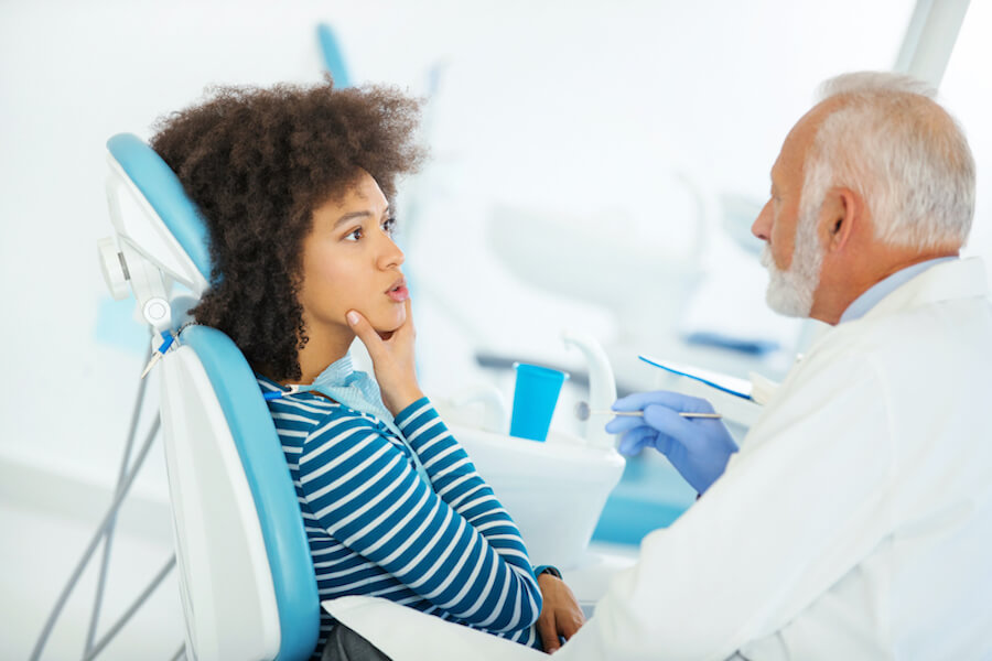 Black woman holds her chin as she sits in a dental chair talking with her dentist