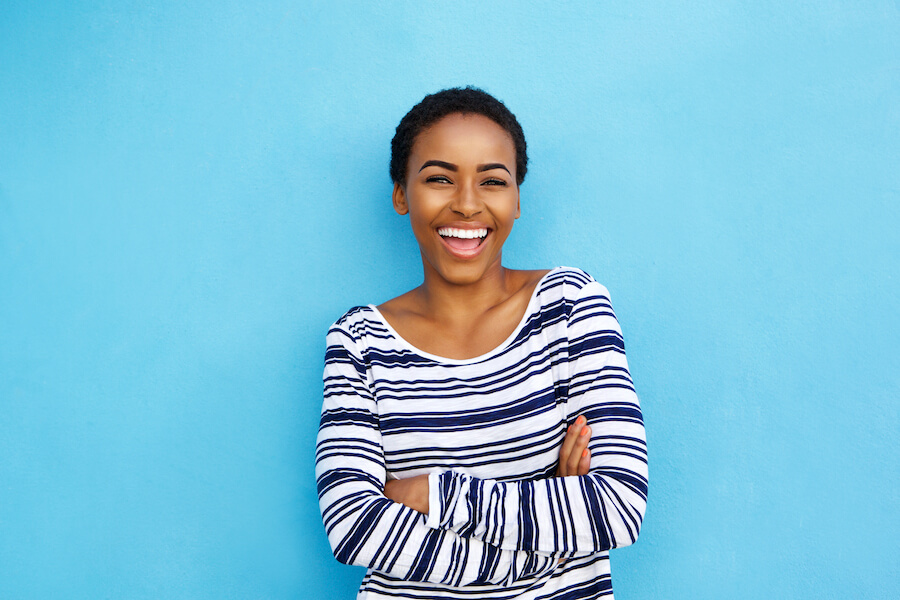 Black woman with strong tooth enamel smiles while standing in a striped shirt against a sky blue wall