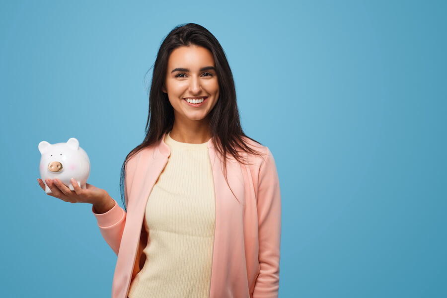 Brunette woman smiles against a blue background while holding a white piggy bank