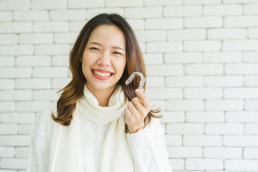 Brunette woman smiles while holding her Invisalign clear aligners while wearing a cream sweater against a white brick wall