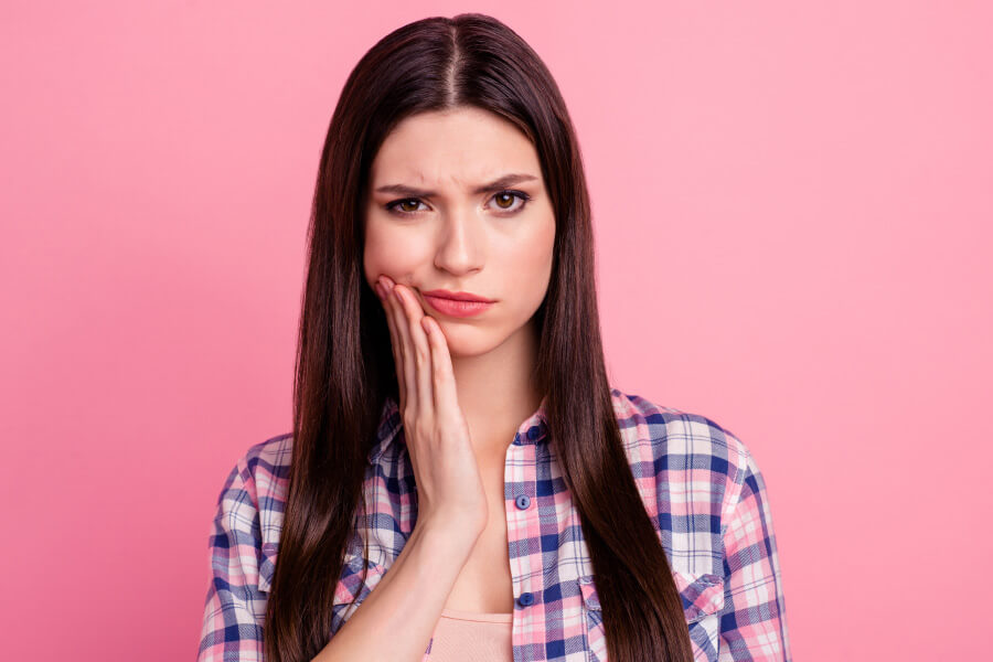 Brunette woman against a pink wall cringes because her teeth feel sensitive to cold