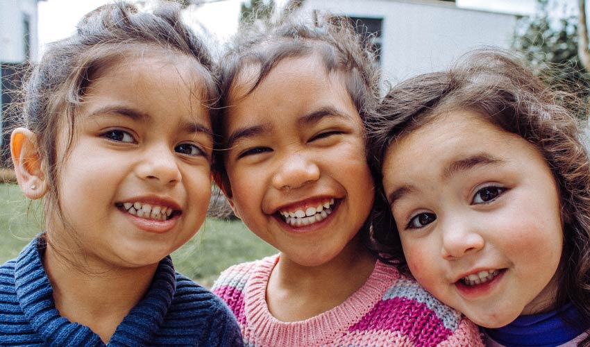 Group of 3 smiling little girls with brown hair standing outside