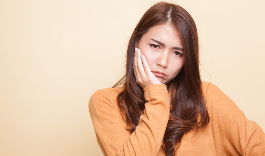 Brunette woman wearing a yellow sweater cringes in pain and touches her cheek due to a toothache